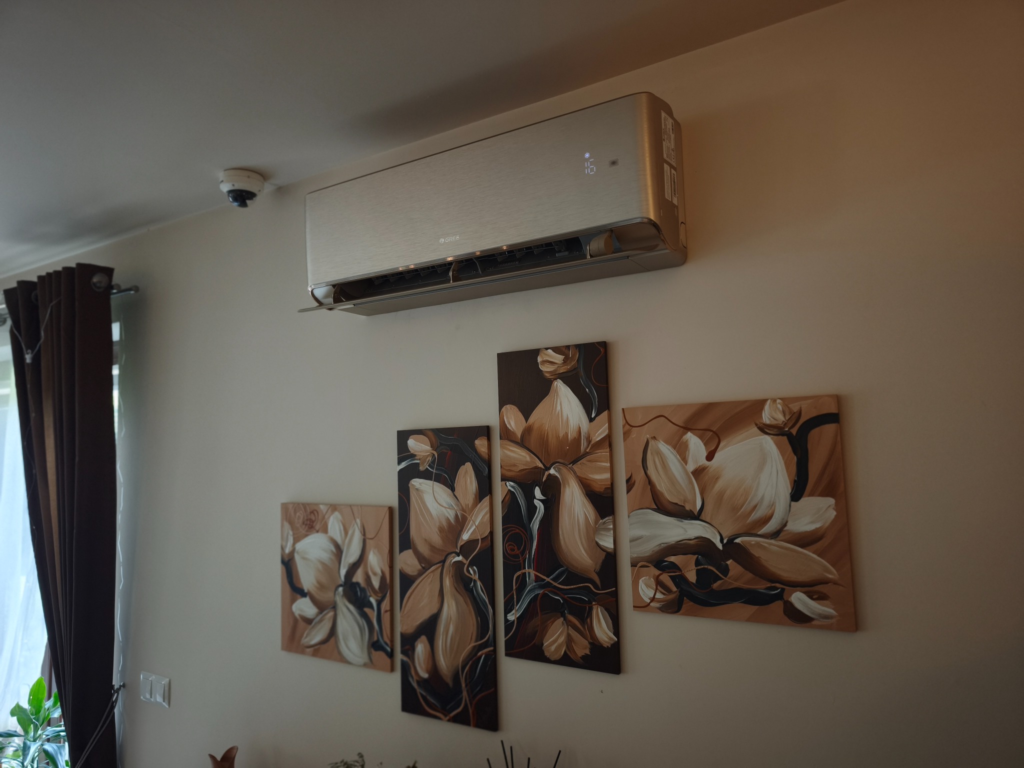 Srebrny klimatyzator ścienny GREE z cyfrowym wyświetlaczem '16' zainstalowany w jasnym pokoju nad czterema beżowo-brązowymi obrazami z motywem magnolii. Widoczna kamera sufitowa.