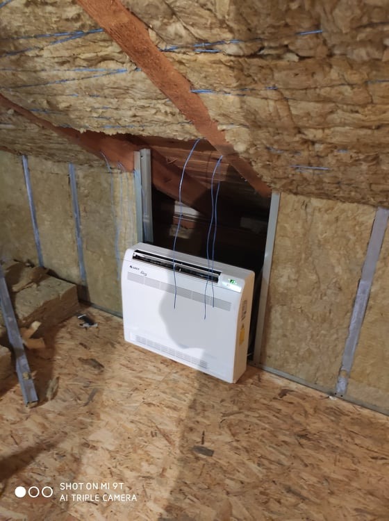 Biała jednostka wewnętrzna klimatyzatora podłogowego marki Gree, umieszczona na poddaszu w trakcie instalacji, widoczne ocieplenie wełną mineralną oraz wystające przewody.