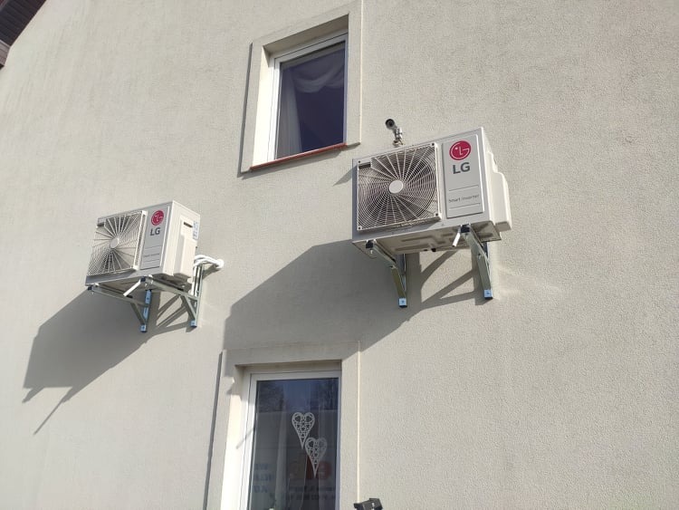 Dwa klimatyzatory LG zamontowane na metalowych wspornikach na jasnej elewacji budynku mieszkalnego, widoczne okna z dekoracjami.