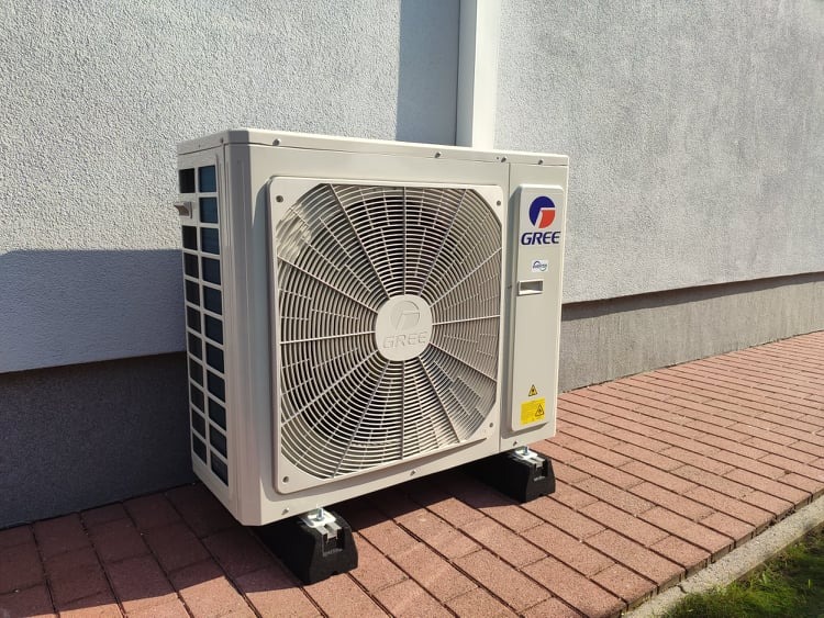 Zewnętrzna jednostka klimatyzacji marki Gree zamontowana na czarnych, gumowych podstawach na tle szarej ściany budynku i czerwonej kostki brukowej.