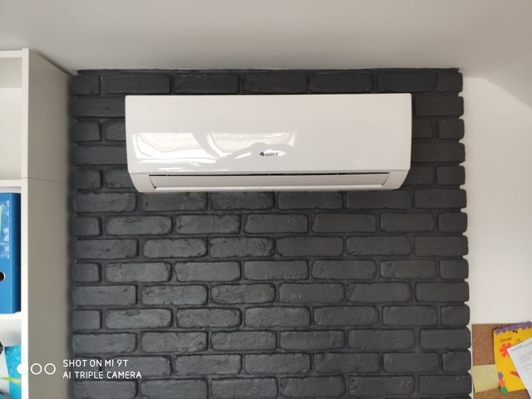 Biała jednostka klimatyzacyjna Gree zamontowana na ścianie z czarnej cegły w pomieszczeniu.