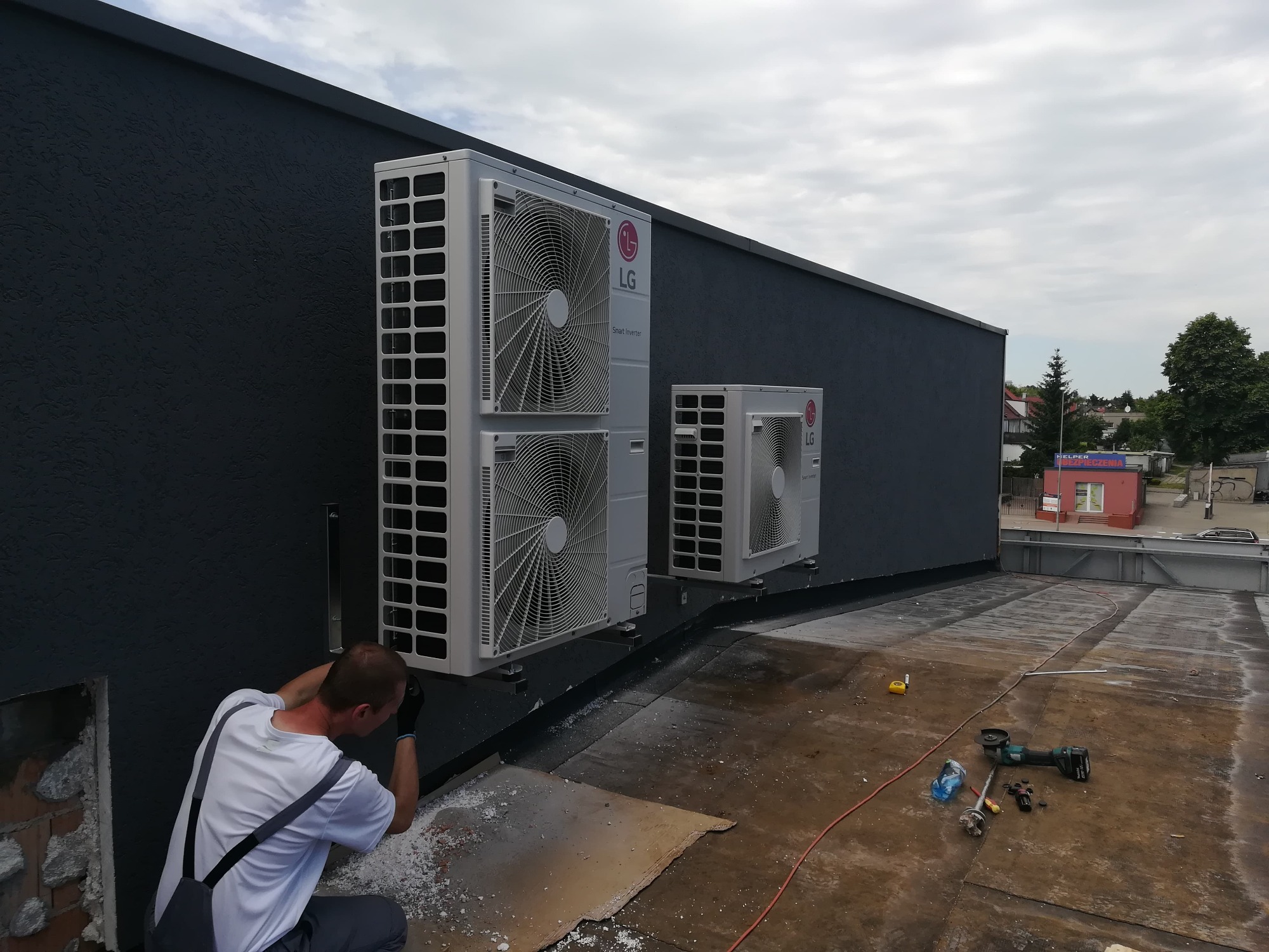 Instalacja dwóch jednostek zewnętrznych klimatyzacji LG na dachu budynku z czarną elewacją, widoczny instalator w trakcie pracy, narzędzia na dachu.
