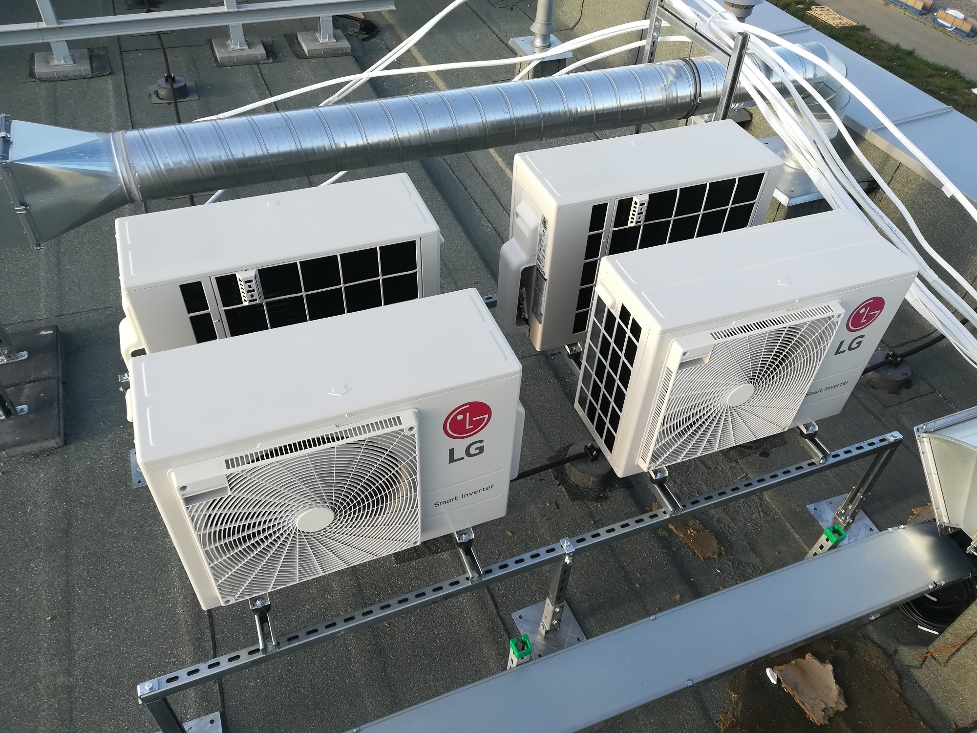 Cztery jednostki zewnętrzne klimatyzacji LG Smart Inverter zamontowane na płaskim dachu, widoczne kanały wentylacyjne i rury instalacyjne.