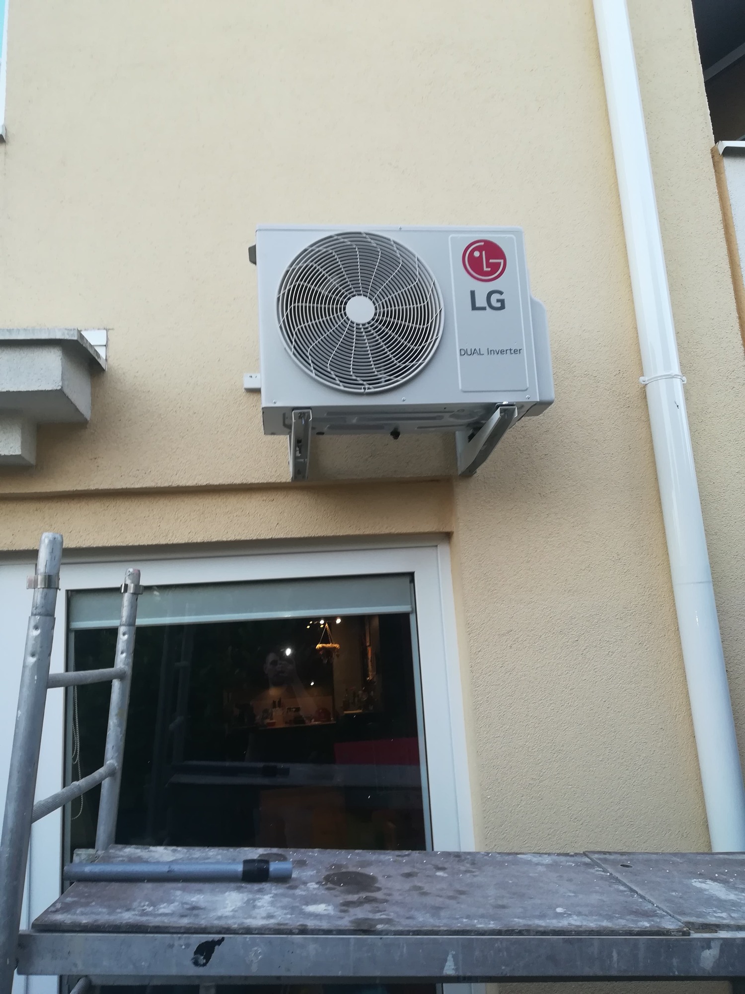 Zewnętrzna jednostka klimatyzacji LG Dual Inverter zamontowana na ścianie budynku, widoczna drabina monterska i odblask twarzy w oknie.