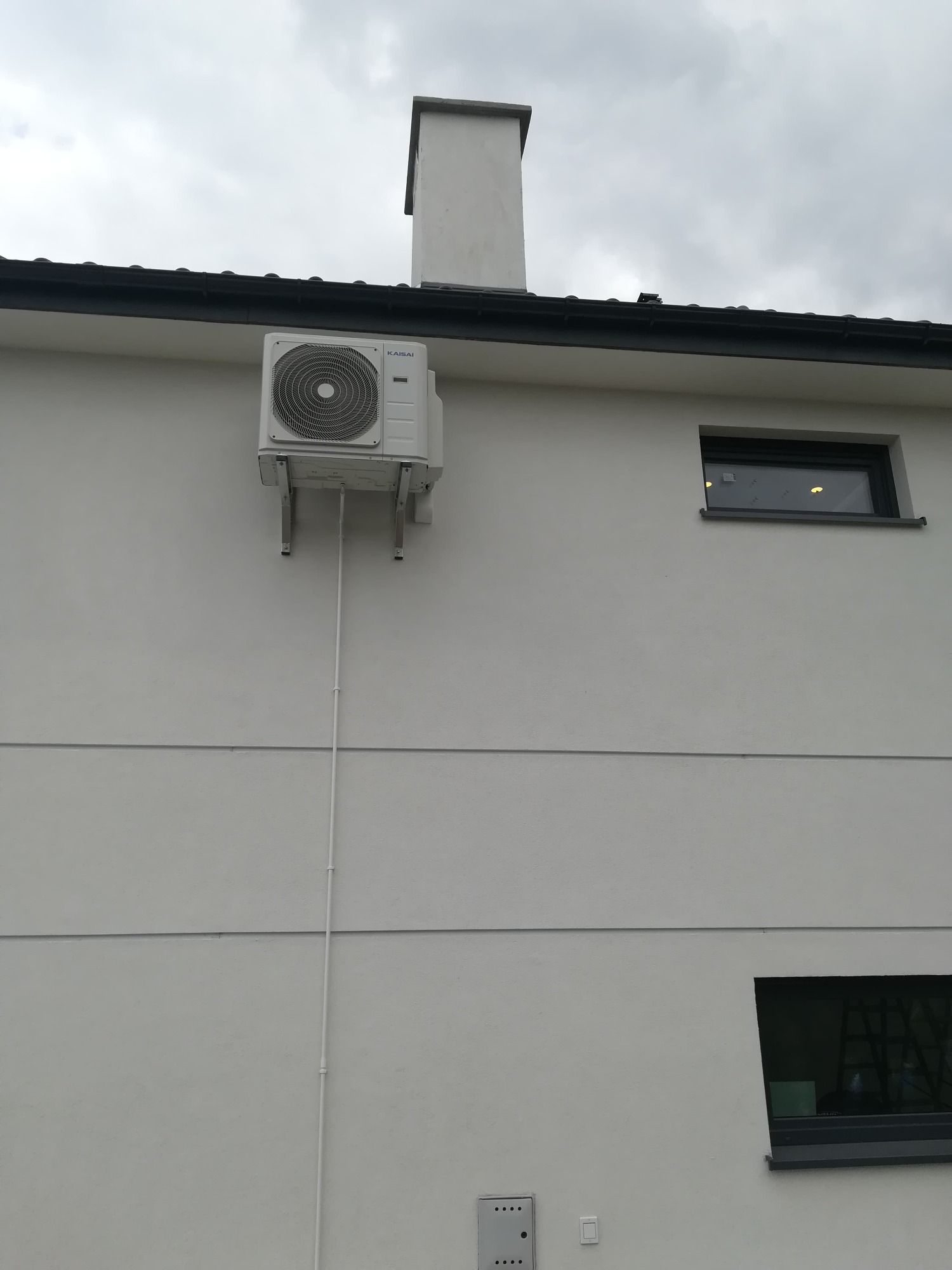Biała jednostka zewnętrzna klimatyzacji Kaisai zamontowana na wspornikach na jasnej elewacji domu, widoczna rura odprowadzająca skropliny, małe okno z czarną ramą w tle.