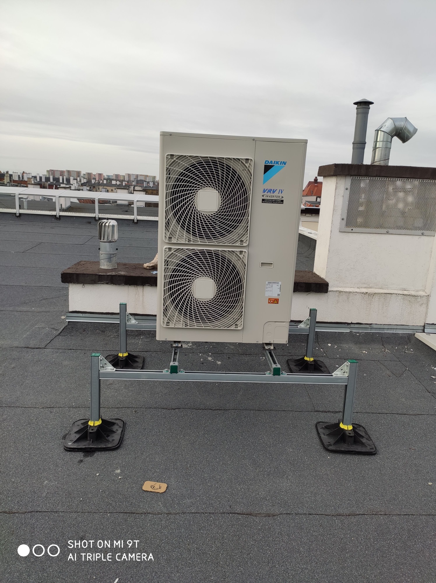 Zewnętrzna jednostka klimatyzacyjna Daikin VRV IV zamontowana na dachu budynku, widoczne elementy montażowe i tło miejskie z kominami wentylacyjnymi.
