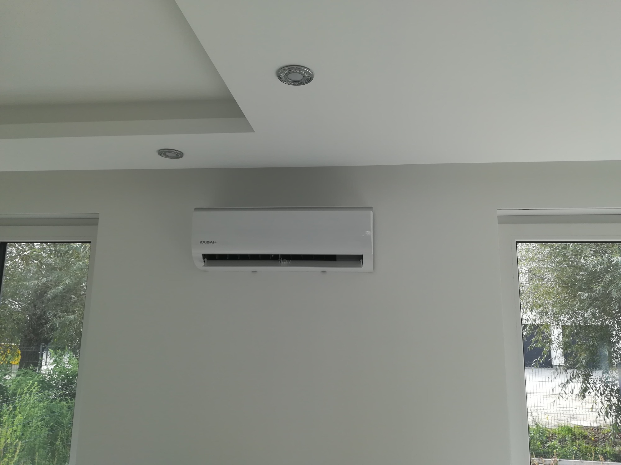 Biała jednostka klimatyzacji Kaisai zamontowana na ścianie w jasnym pomieszczeniu, widok z dołu na wprost, okno z widokiem na zieleń w tle.