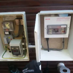 Dwa otwarte białe skrzynki elektryczne na brązowej ścianie, w jednej licznik i bezpieczniki, w drugiej tylko bezpieczniki, widoczna dłoń w czarnej rękawicy z logiem.