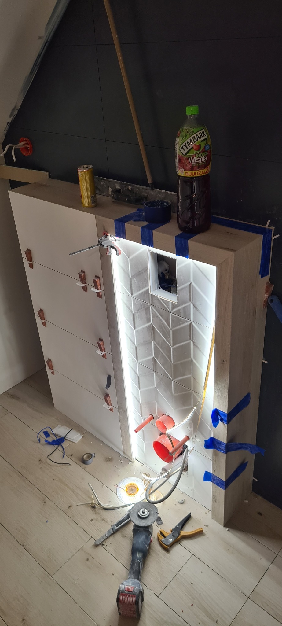 Montaż podtynkowej toalety z podświetleniem LED i wzorem w jodełkę, widoczne narzędzia, klej, puszka napoju i sok na blacie, w trakcie prac wykończeniowych.