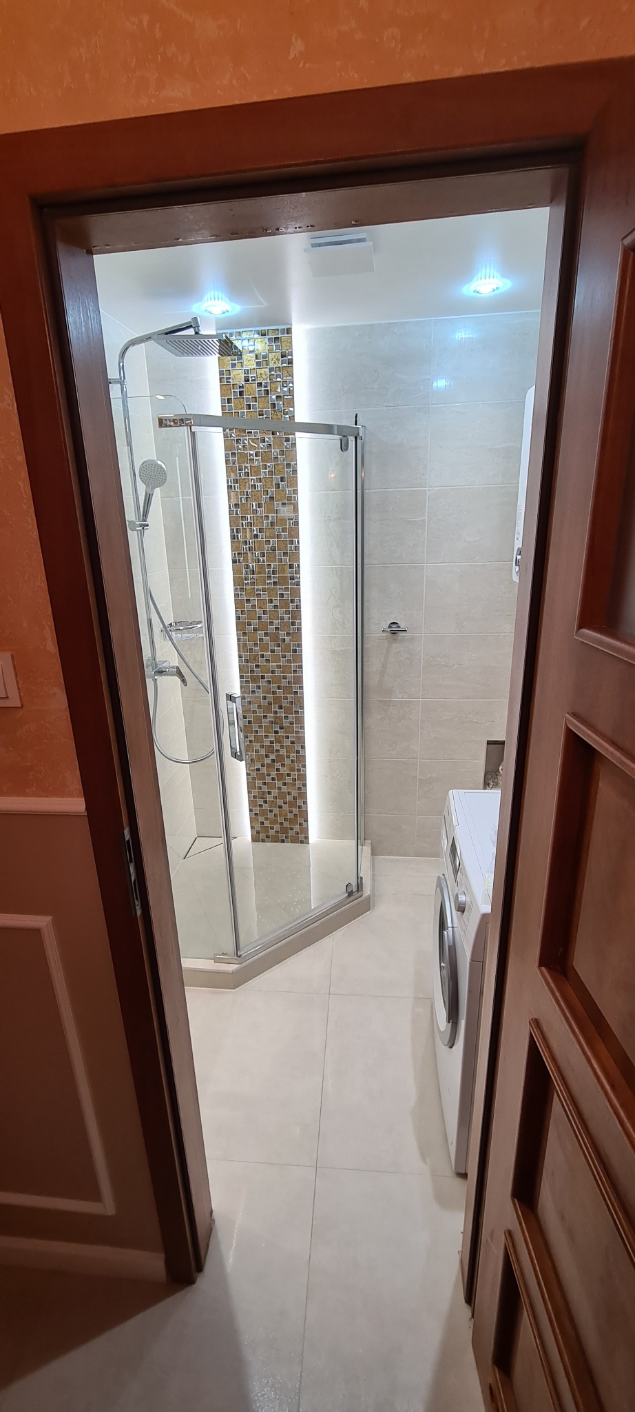 Nowoczesna kabina prysznicowa z mozaiką i deszczownicą, widok zza drewnianych drzwi, obok pralka w łazience wykończonej jasnymi płytkami.