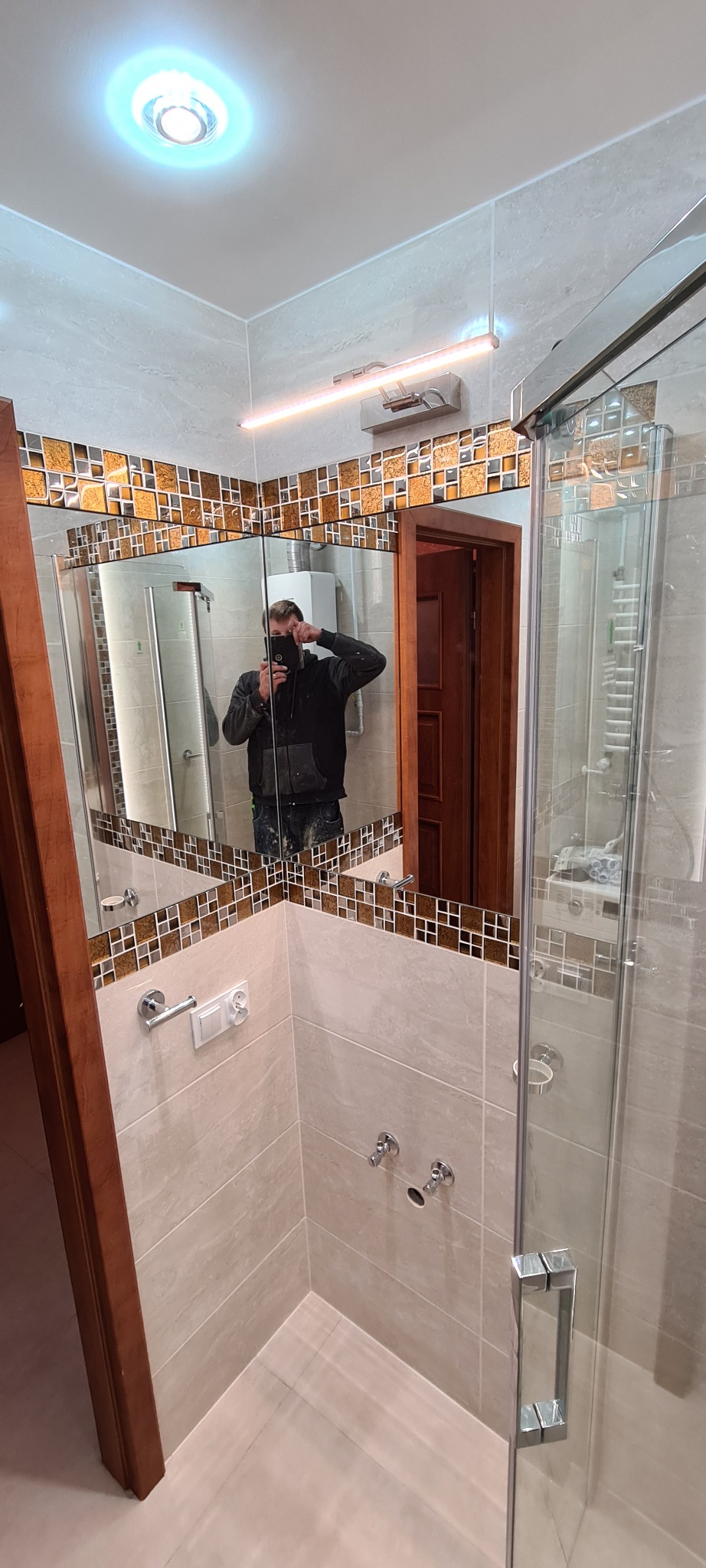 Wykończona łazienka z beżowymi płytkami, mozaiką w odcieniach brązu i szklaną kabiną prysznicową, odbicie fotografa w lustrze.