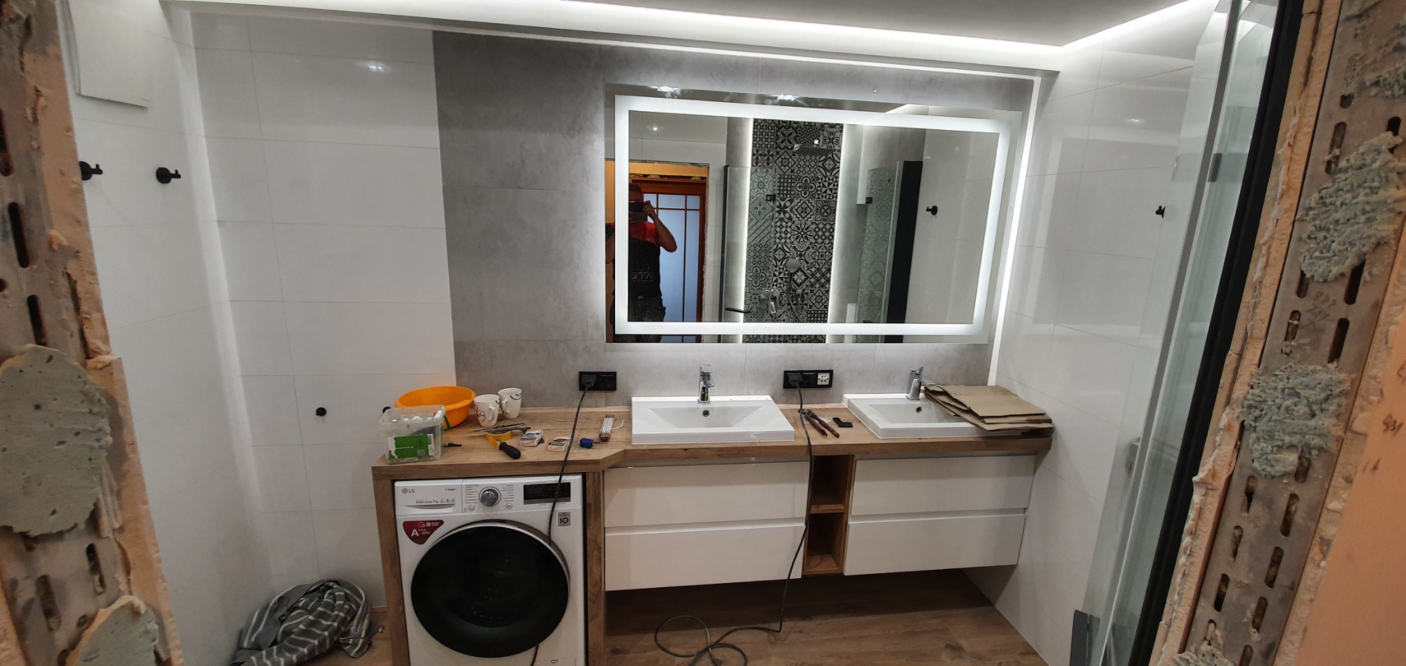 Remontowana łazienka w Gdyni z podwójną umywalką, pralką i dużym lustrem z podświetleniem LED, widoczne narzędzia i materiały budowlane sugerują trwające prace wykończeniowe.