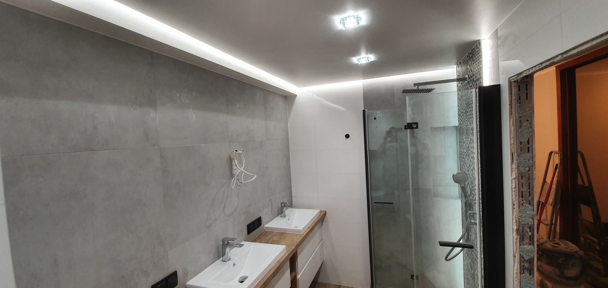 Nowoczesna łazienka z dwiema umywalkami na drewnianym blacie, kabina prysznicowa z czarnymi elementami, oświetlenie LED podsufitowe, szare płytki na ścianach, widoczny fragment niedokończonego...