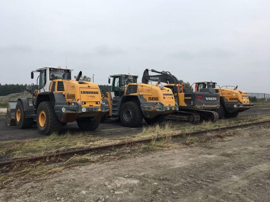Rząd żółtych maszyn budowlanych Liebherr i Volvo, w tym ładowarki kołowe i koparka gąsienicowa, stojących na placu budowy z torami kolejowymi i hałdą ziemi w tle.