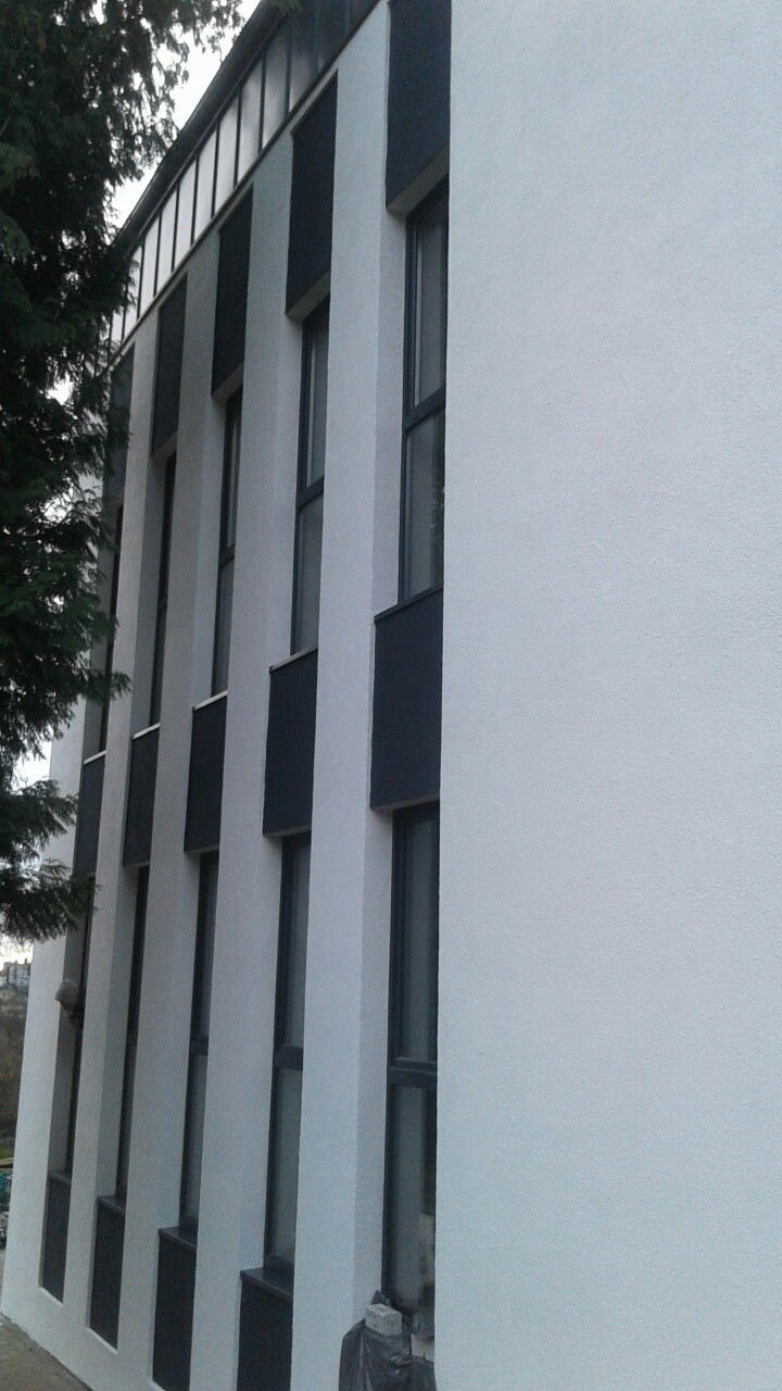 Boczny widok budynku po zakończonym ociepleniu elewacji, z widocznymi oknami w ciemnych obramowaniach i świeżo pomalowaną białą elewacją, z fragmentem drzewa po lewej stronie.