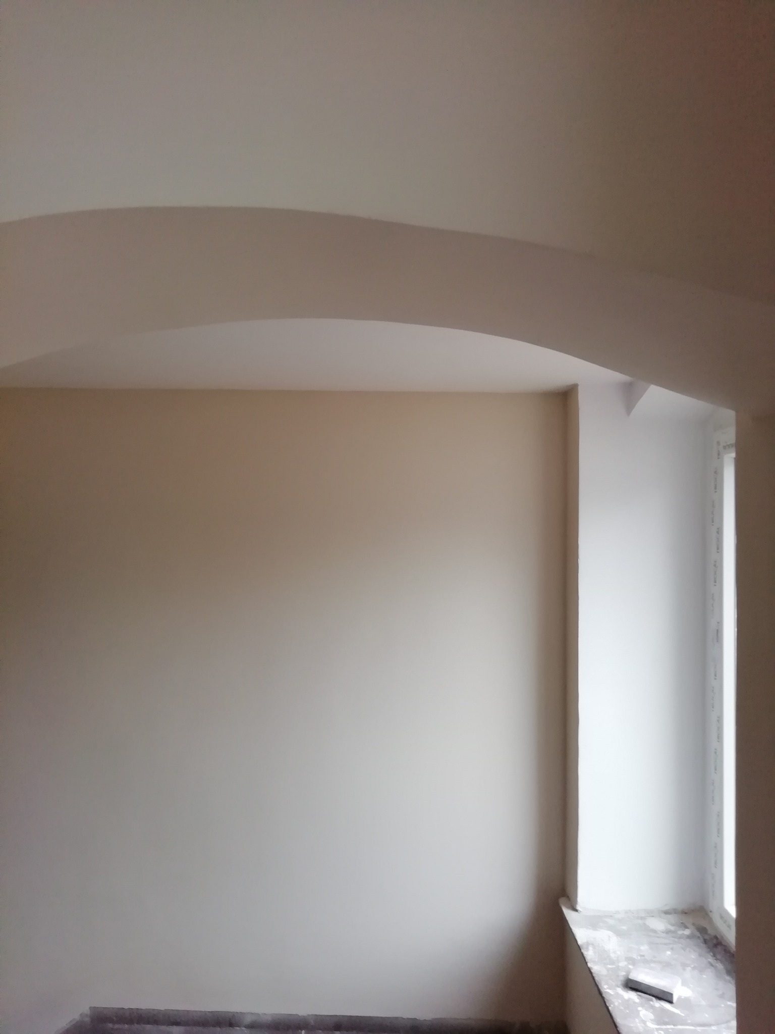 Wnętrze pokoju po świeżym pomalowaniu na beżowo, z zaokrąglonym przejściem ściany w sufit i widocznym oknem z parapetem, na którym leży szpachelka.