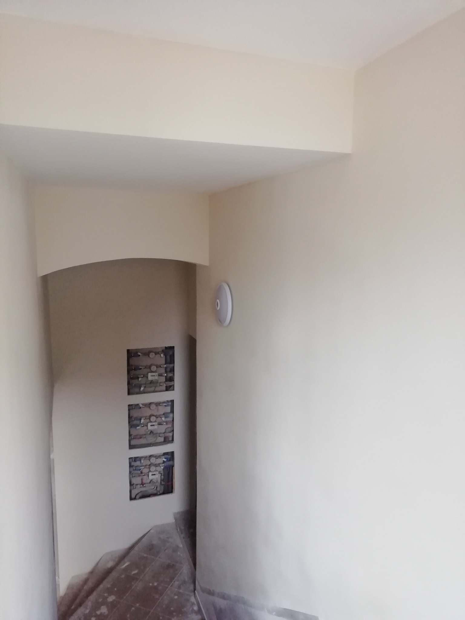 Świeżo pomalowane na beżowo ściany korytarza z widocznymi otworami rewizyjnymi na instalacje oraz lampą sufitową. Podłoga częściowo zabezpieczona folią malarską.