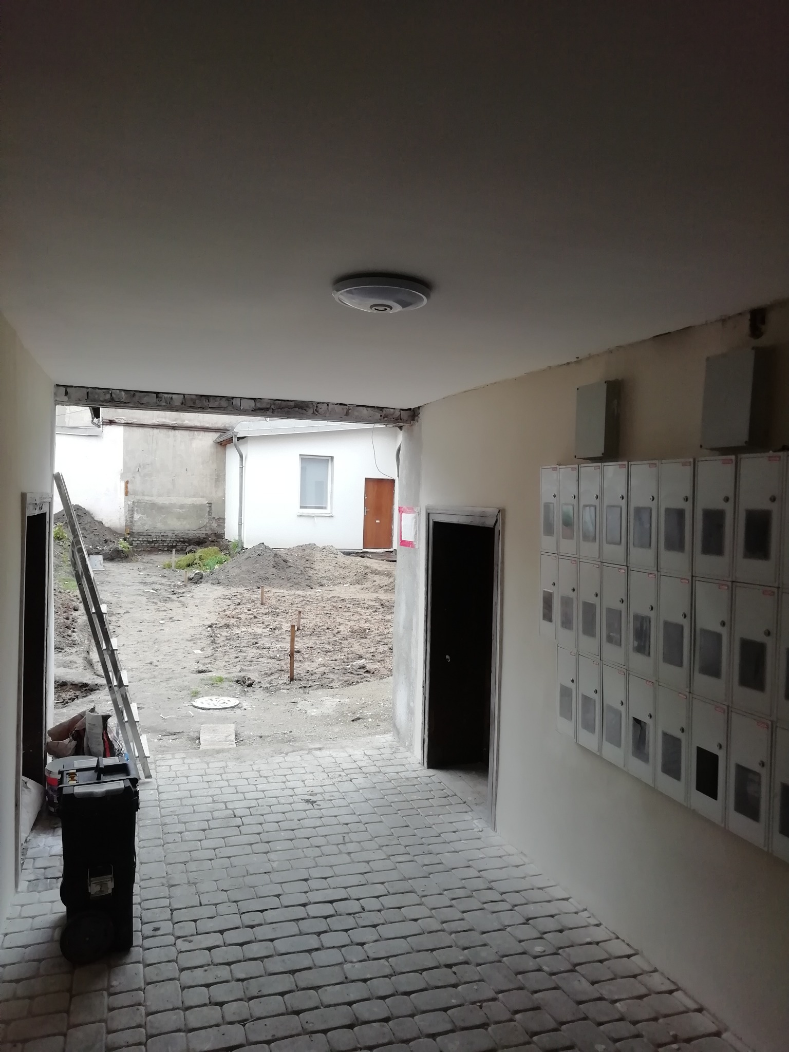 Korytarz w trakcie remontu z widoczną drabiną, narzędziami i stertami ziemi na zewnątrz, ukazujący etap prac przygotowawczych do wykończenia elewacji.
