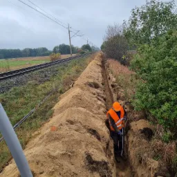 Pracownik w odblaskowej kamizelce kopie wąski rów w ziemi wzdłuż torów kolejowych, prawdopodobnie pod kable; widoczna wykopana ziemia i fragment drzewa.