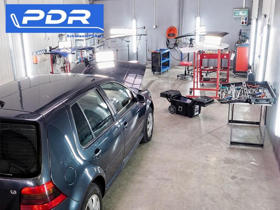 Samochód z otwartą maską w warsztacie PDR, narzędzia do bezinwazyjnego usuwania wgnieceń na stole, oświetlenie warsztatowe.
