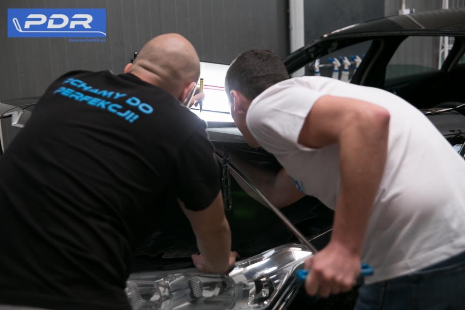 Dwóch mężczyzn pochylonych nad czarnym samochodem, analizujących wgniecenie przy użyciu specjalistycznego oświetlenia i narzędzi PDR; jeden z nich ma na koszulce napis 'Dążymy do perfekcji!'.