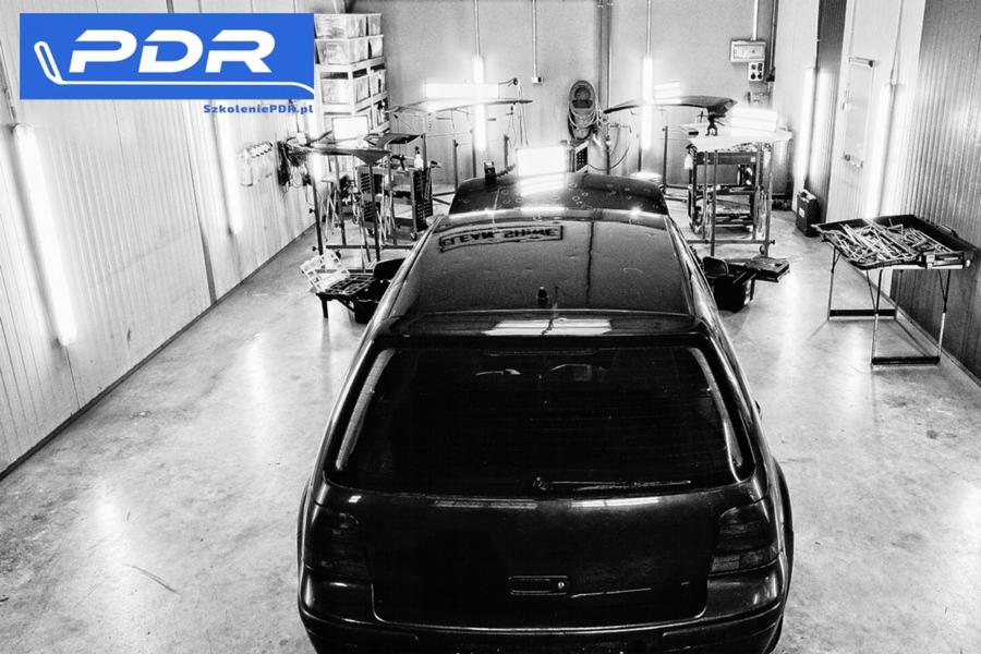 Czarno-białe zdjęcie tylnej części czarnego samochodu w warsztacie PDR (Paintless Dent Repair) z widocznym logo firmy PDR i narzędziami do naprawy wgnieceń bez lakierowania.