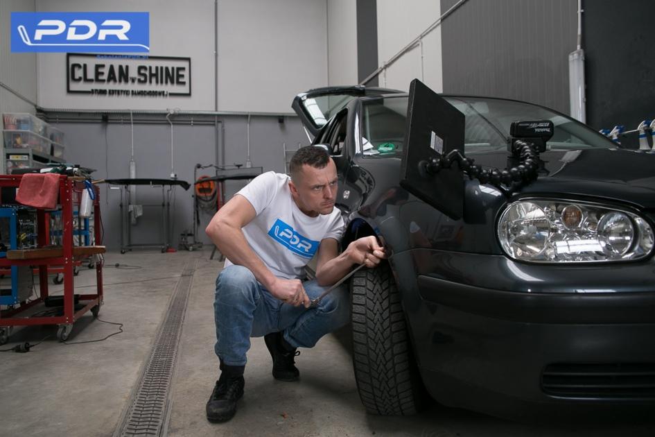 Technik PDR usuwa wgniecenie na błotniku czarnego samochodu za pomocą narzędzia dźwigniowego, z przyssawką i lampą inspekcyjną na masce.