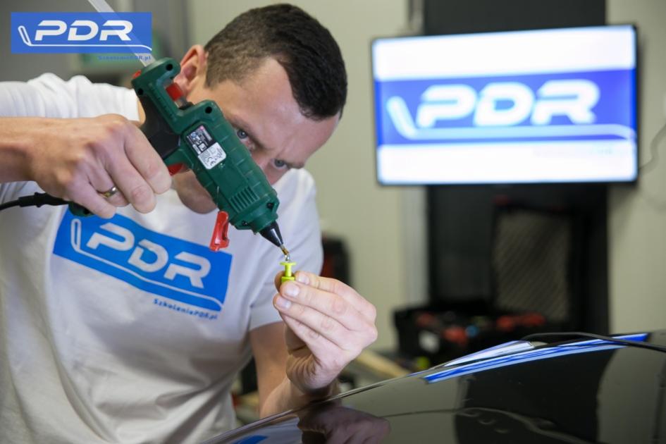 Mężczyzna z pistoletem do kleju, przygotowujący element do naprawy karoserii metodą PDR, w tle monitor z logo PDR.