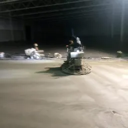 Pracownik w kombinezonie wygładza świeżo wylaną betonową podłogę w hali za pomocą zacieraczki mechanicznej; widoczne ślady butów na powierzchni.