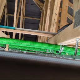 Zielone, karbowane przewody wentylacyjne zamocowane do drewnianej konstrukcji poddasza z widocznymi metalowymi uchwytami i elementami mocującymi.