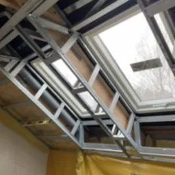 Stelaż metalowy pod zabudowę okna dachowego na poddaszu, widoczne płyty OSB i izolacja folią w kolorze żółtym, częściowo zamontowana rura wentylacyjna.