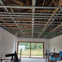 Metalowa konstrukcja sufitowa z widocznymi elementami drewnianymi i instalacjami, przygotowana do montażu płyt, widok z wnętrza pomieszczenia z dużym oknem i rusztowaniami roboczymi.