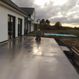 Układanie jasnoszarych płytek na tarasie domu jednorodzinnego; pracownik w kombinezonie klęczy na powierzchni, w tle widoczne niebo z chmurami i zarys krajobrazu.