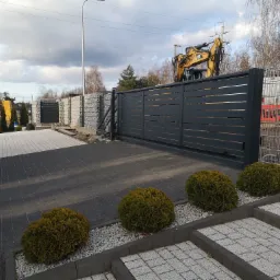 Nowoczesna, przesuwna brama wjazdowa z poziomymi panelami, widoczna z zewnątrz posesji. W tle koparka CAT oraz ogrodzenie z siatki. Podłoże wyłożone kostką brukową, ozdobione kulistymi tujami...