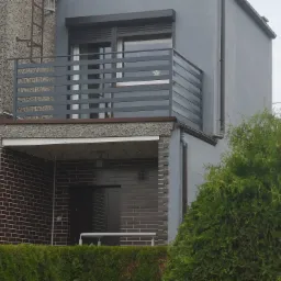 Szara, pozioma balustrada balkonowa z metalowych profili na tle szarej elewacji dwukondygnacyjnego domu z ceglanym wykończeniem i zielonym żywopłotem.