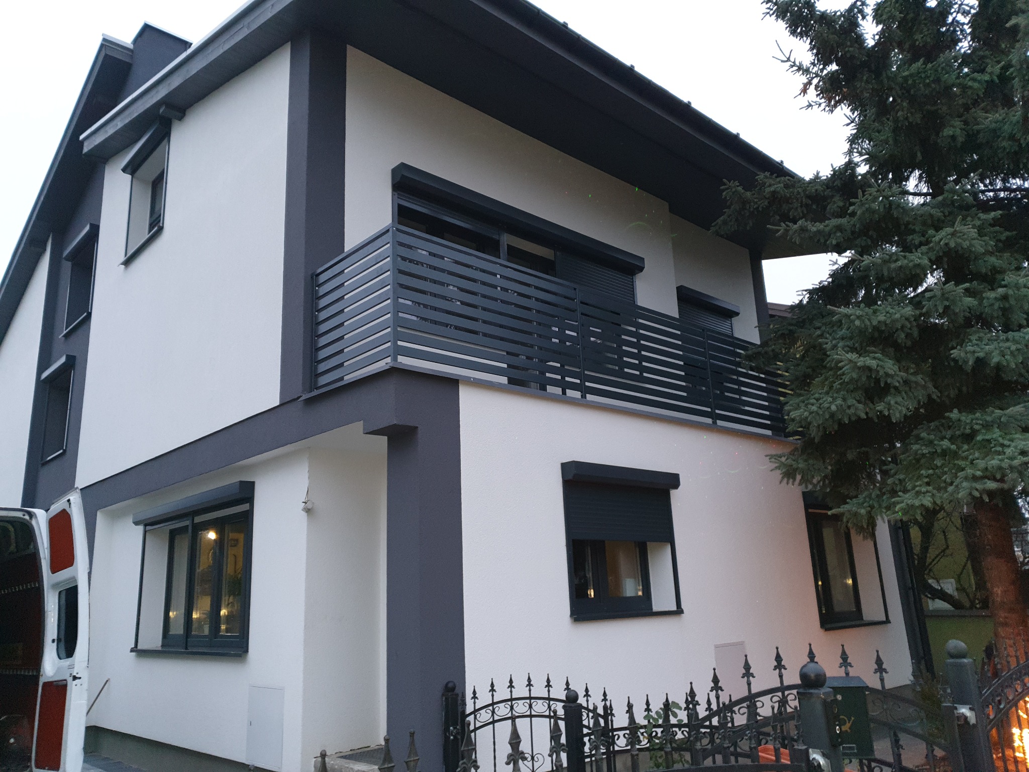 Dwupiętrowy dom z szarą, poziomą balustradą na balkonie, szarymi ramami okiennymi i roletami zewnętrznymi, widziany z poziomu ulicy przez ozdobne, metalowe ogrodzenie.