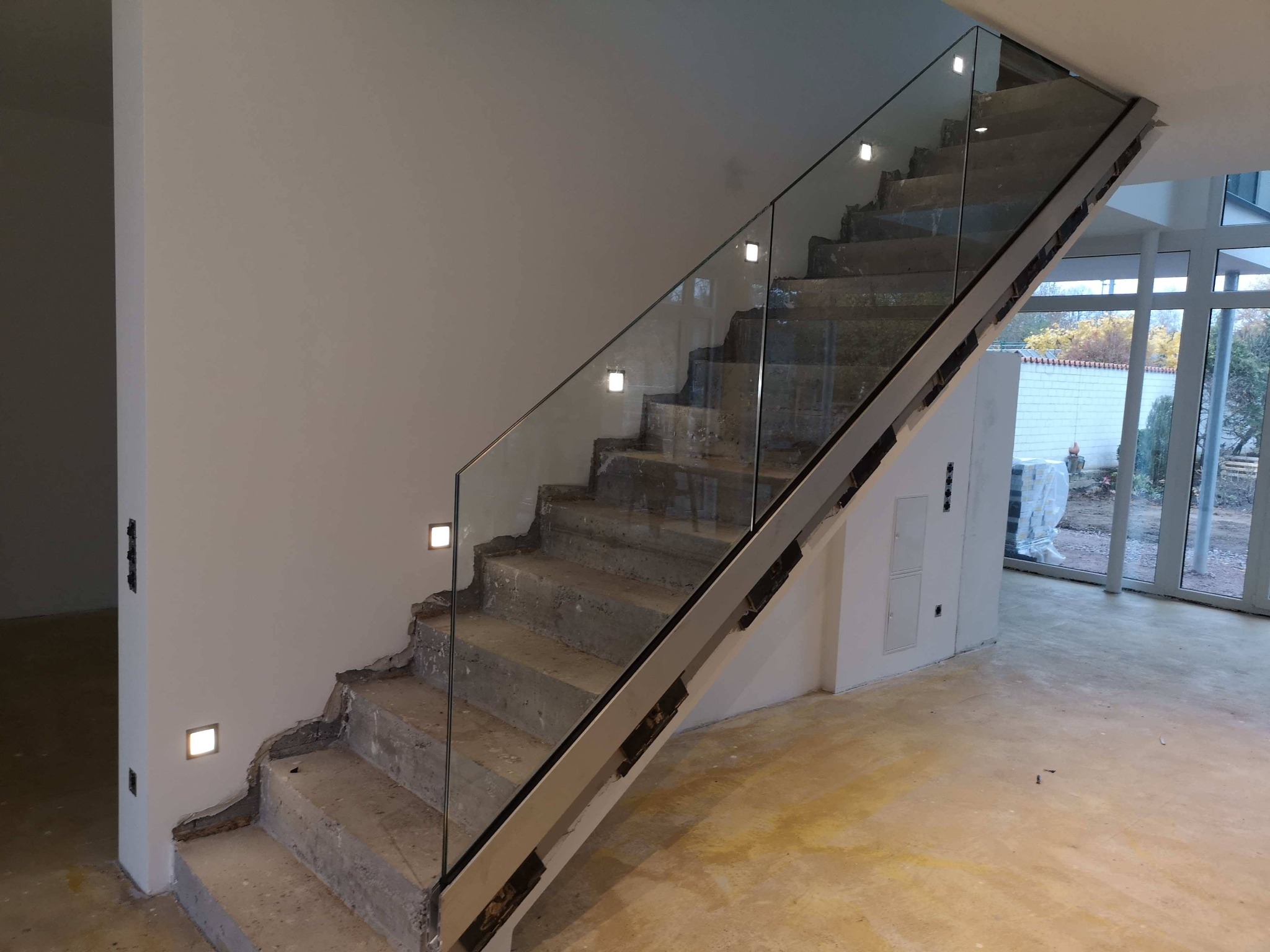 Wewnętrzne schody betonowe z zamontowaną szklaną balustradą z podświetleniem LED w ścianie, widoczne surowe wykończenie ścian i podłogi.