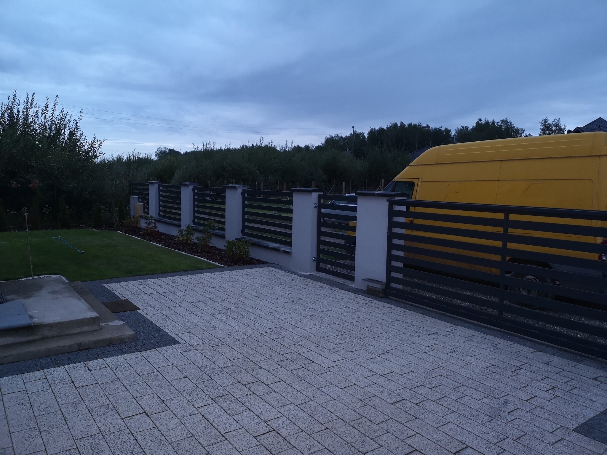 Nowoczesne ogrodzenie posesji z poziomymi przęsłami i murowanymi słupkami, widoczne z perspektywy podjazdu wyłożonego kostką brukową, z żółtym samochodem dostawczym zaparkowanym za ogrodzeniem.