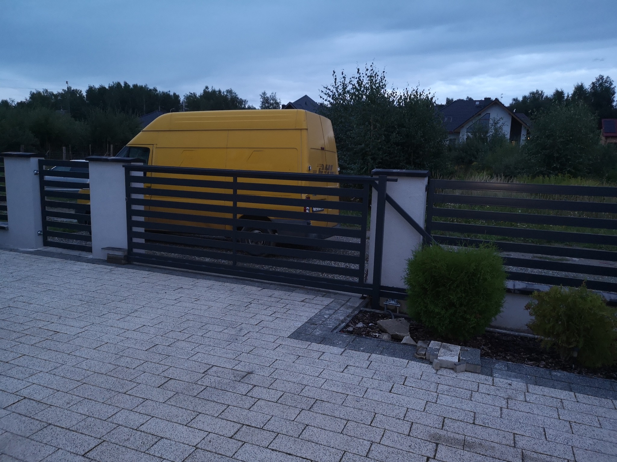 Nowoczesna przesuwna brama wjazdowa z poziomych profili, widoczna z żółtym samochodem dostawczym w tle, ułożona kostka brukowa na podjeździe.