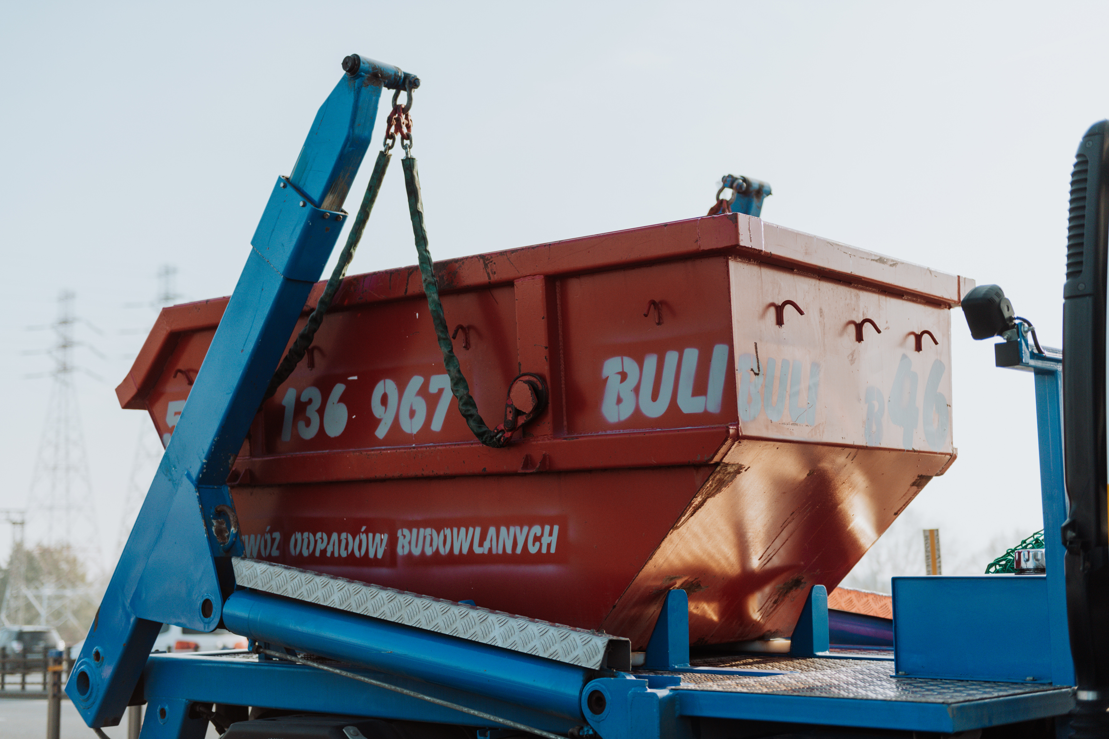 Czerwony kontener na odpady budowlane z numerem telefonu, podnoszony przez niebieski dźwig na tle jasnego nieba i częściowo widocznych drzew.