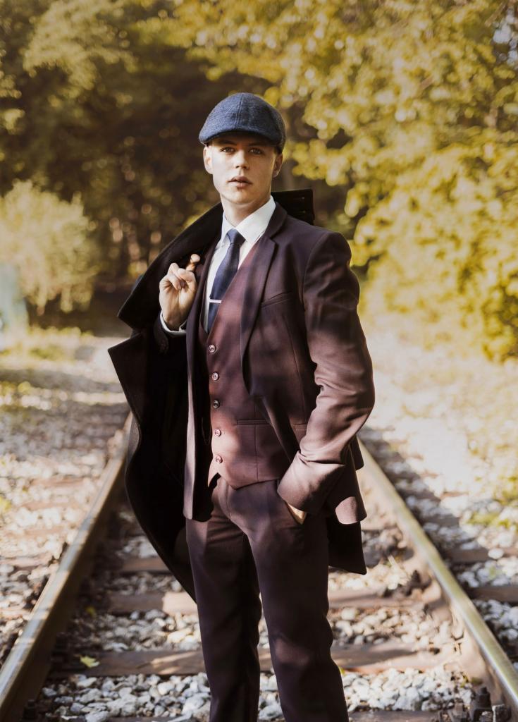 Młody mężczyzna w brązowym garniturze i kaszkiecie stoi na torach kolejowych, trzymając płaszcz na ramieniu.