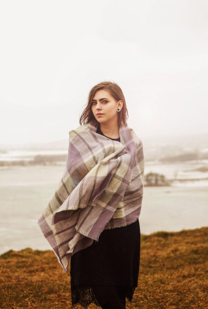 Portret kobiety w Zielonej Górze, owiniętej w duży, pastelowy pled w kratę na tle mglistego krajobrazu.