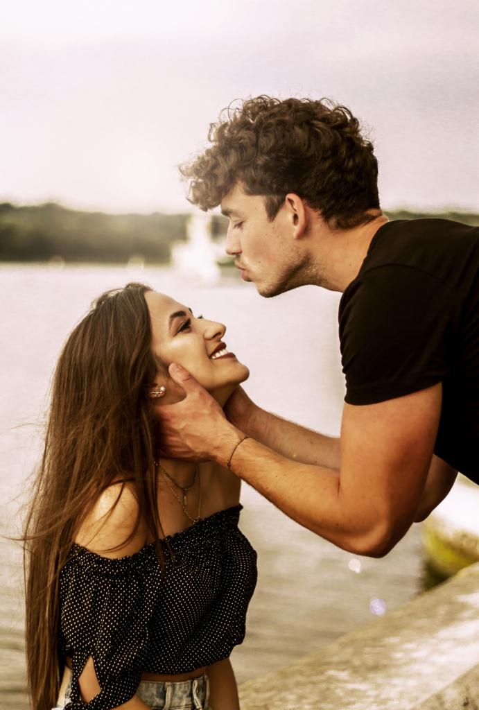 Młoda para: on z kręconymi włosami delikatnie dotyka jej twarzy, ona uśmiecha się, ubrana w bluzkę w groszki, w tle woda i zieleń.