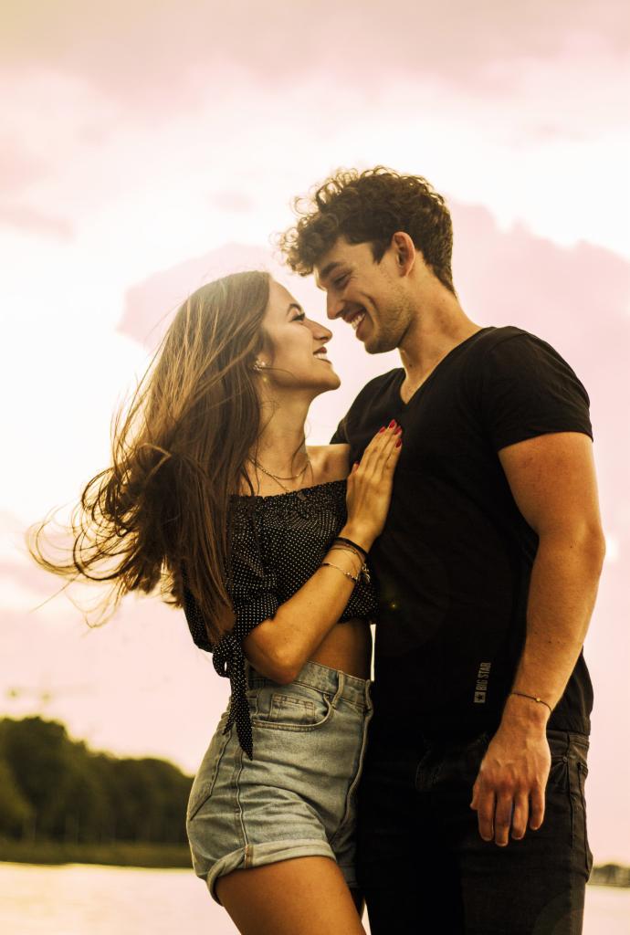 Uśmiechnięta para zakochanych patrzy na siebie na tle różowego nieba o zachodzie słońca; ona w bluzce w kropki i jeansowych szortach, on w czarnym T-shircie.