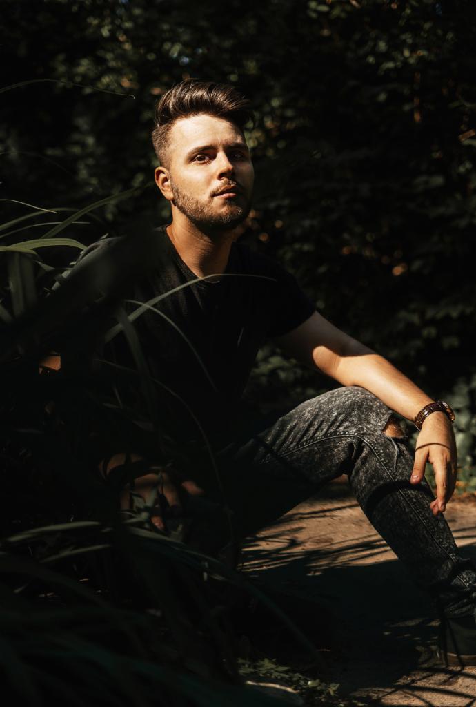 Młody mężczyzna z brodą, ubrany w czarną koszulkę i dżinsy z przetarciami, siedzi na ziemi w otoczeniu zieleni, spoglądając w górę w zamyśleniu.