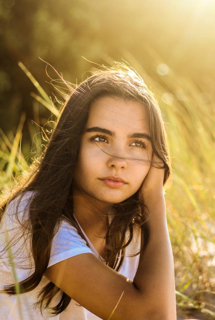 Portret młodej kobiety z długimi brązowymi włosami, opierającej głowę na dłoni, w otoczeniu wysokich traw w złotym świetle zachodzącego słońca.