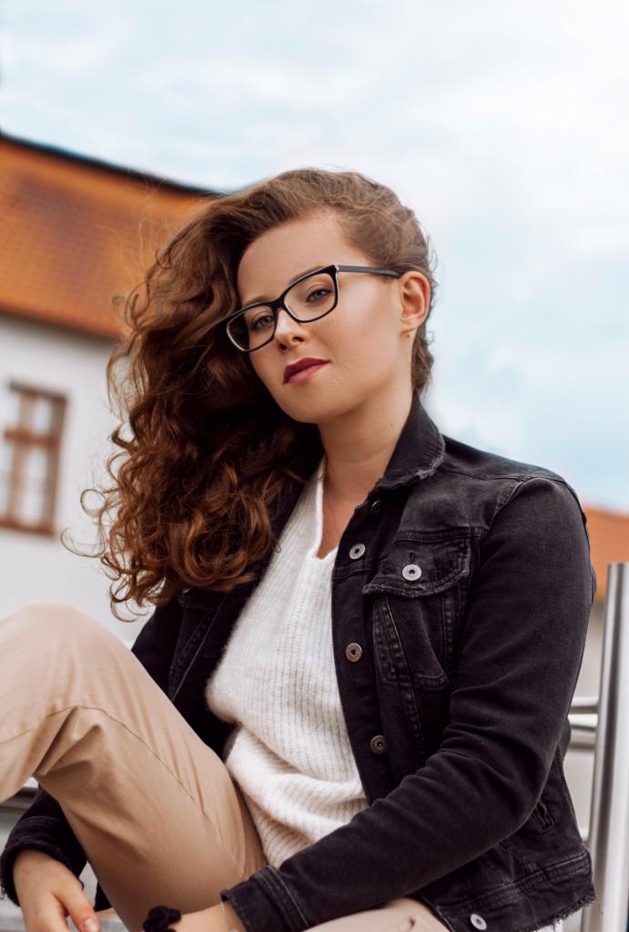 Portret kobiety w okularach i jeansowej kurtce, siedzącej na tle budynku z czerwoną dachówką.