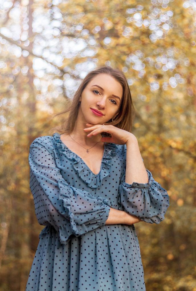 Portret młodej kobiety w błękitnej sukience w groszki, opierającej podbródek na dłoni na tle jesiennych drzew z rozmytym tłem.