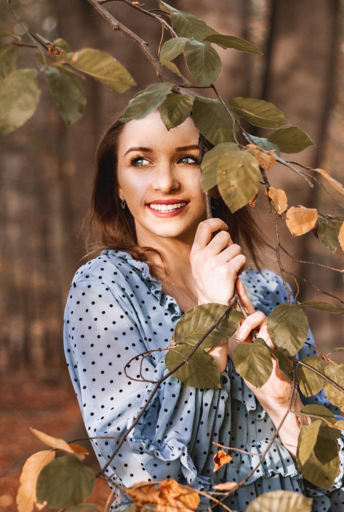 Uśmiechnięta młoda kobieta w niebieskiej sukience w kropki, pozująca w lesie, trzymająca gałązkę z liśćmi, z delikatnym oświetleniem i rozmyciem tła.