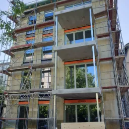 Budynek w trakcie montażu okien z widocznym rusztowaniem, oknami z logo 'roma' i stertą drewna u podnóża.